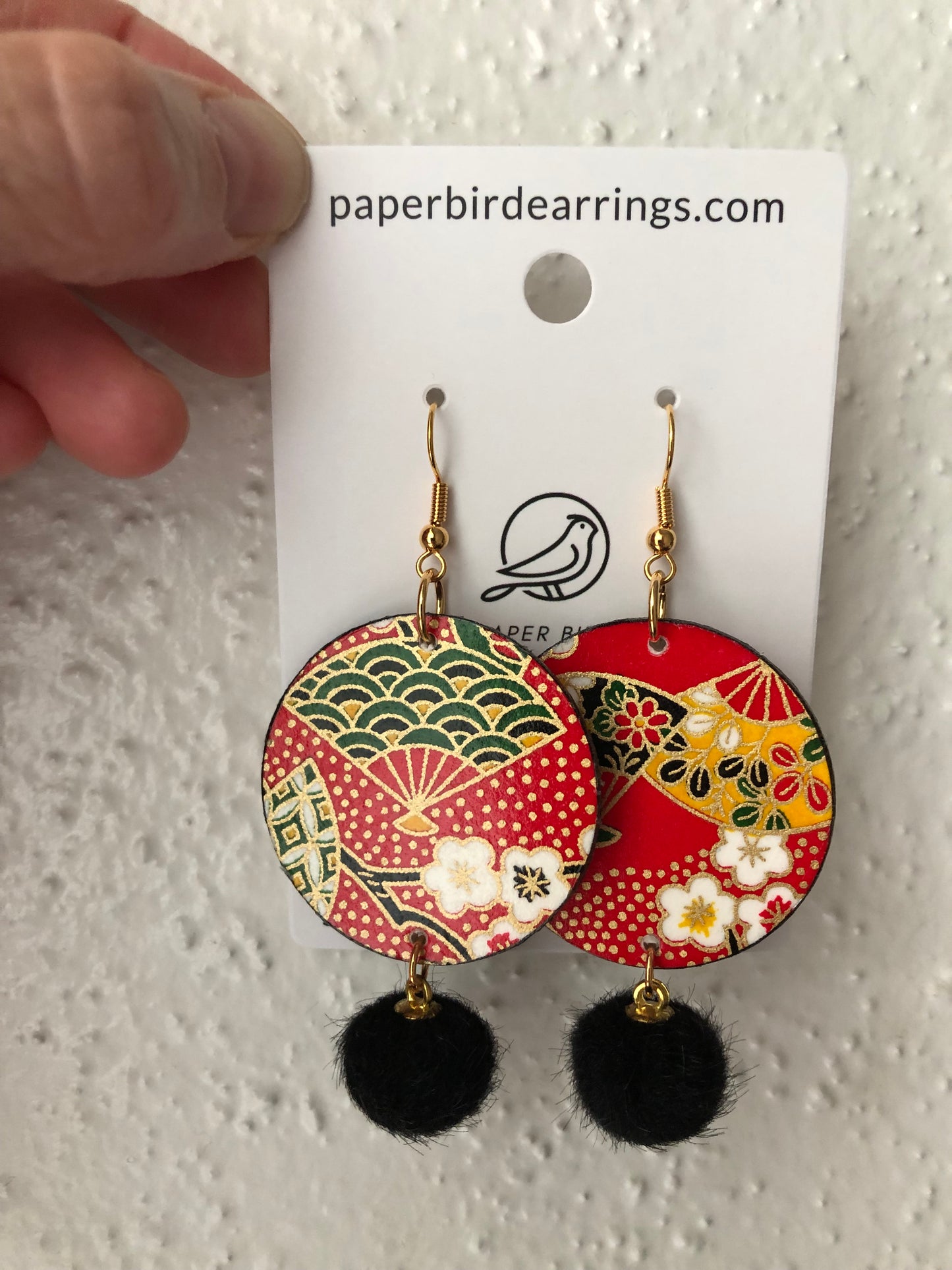 handmade paper earrings of patterned folding fans on a red background design with black pom-poms