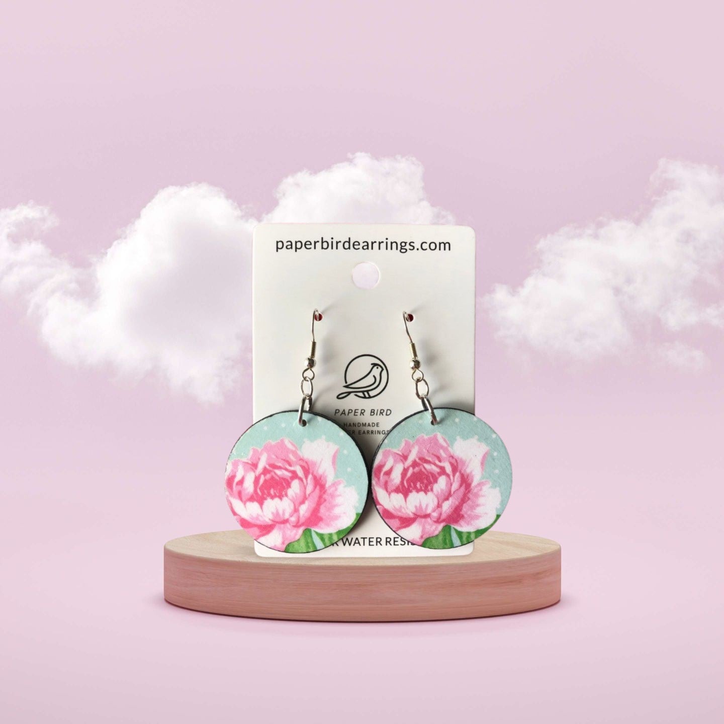 Floral earrings on a wooden stand with a branded card against a soft pink background