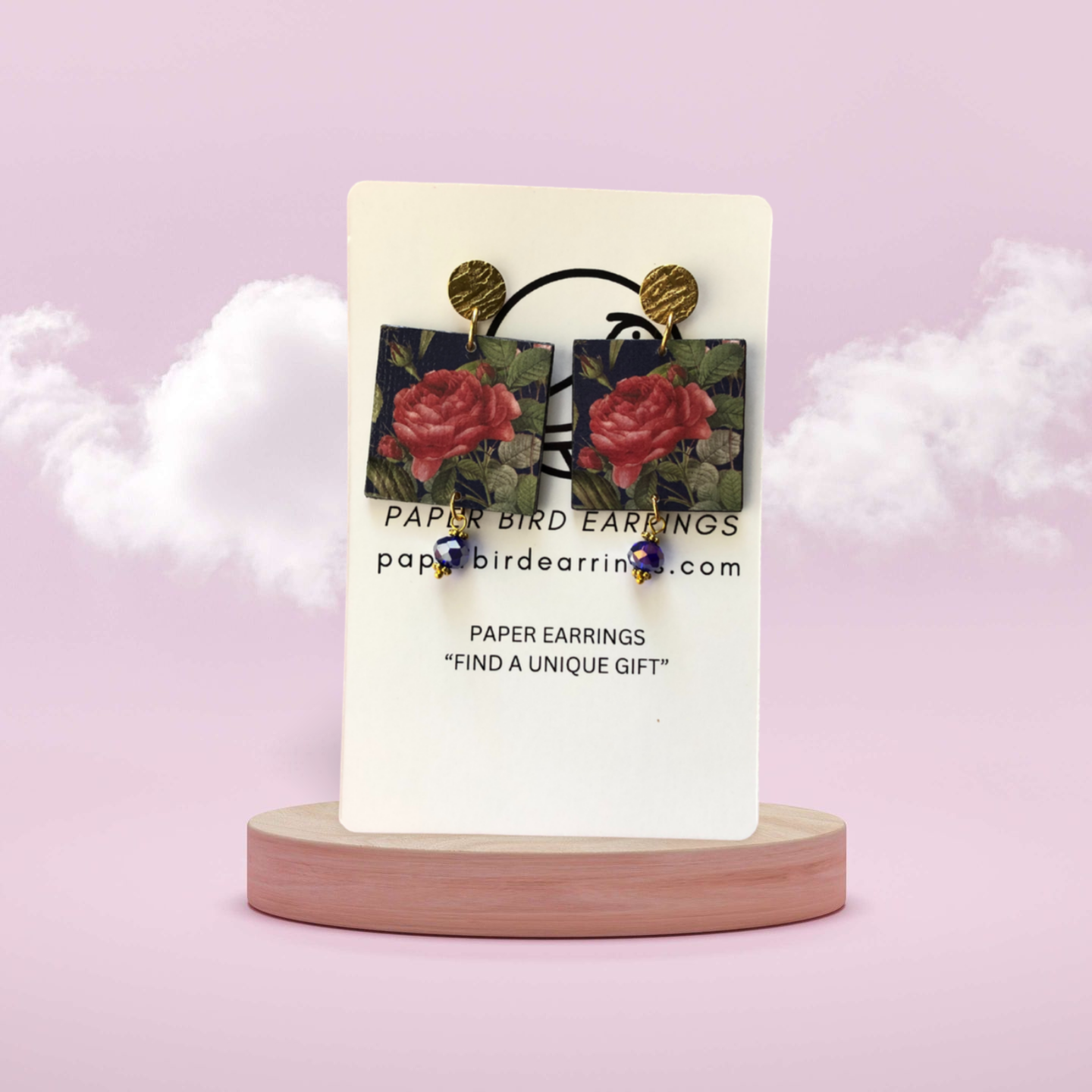 A pair of paper earrings with a rose flower design on a blue background, displayed on a wooden stand with the brand name 'PAPER BIRD EARRINGS' above.
