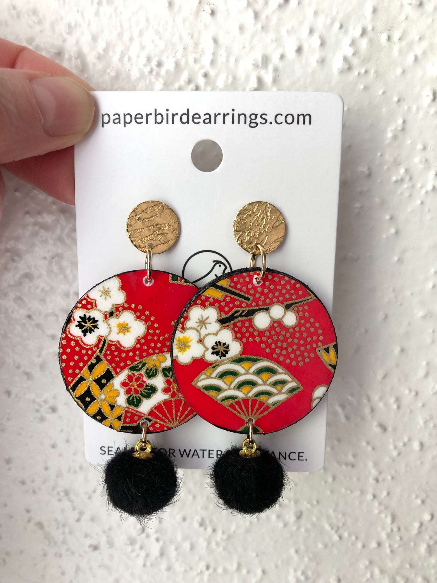 Decorative paper earrings with floral and geometric design on red background with black pom poms 