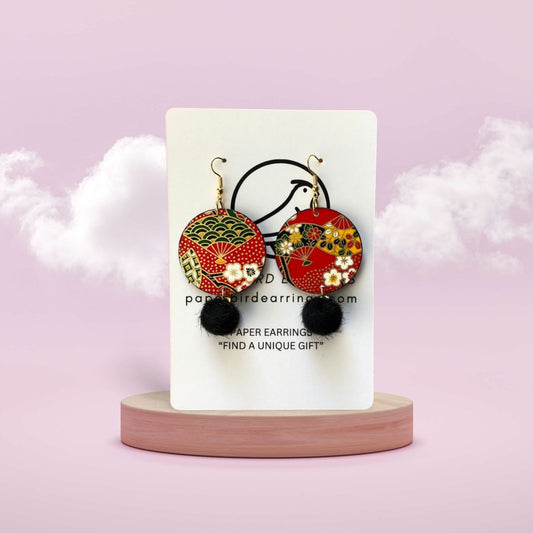 Pair of colorful earrings with pom-pom ends on a white card against a pink background