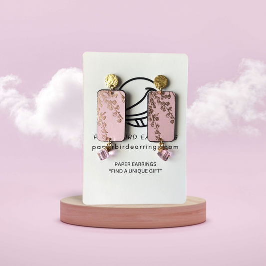 A pair of gold flower on pink rectangle earrings displayed on a wooden stand with a cloud and sky backdrop.