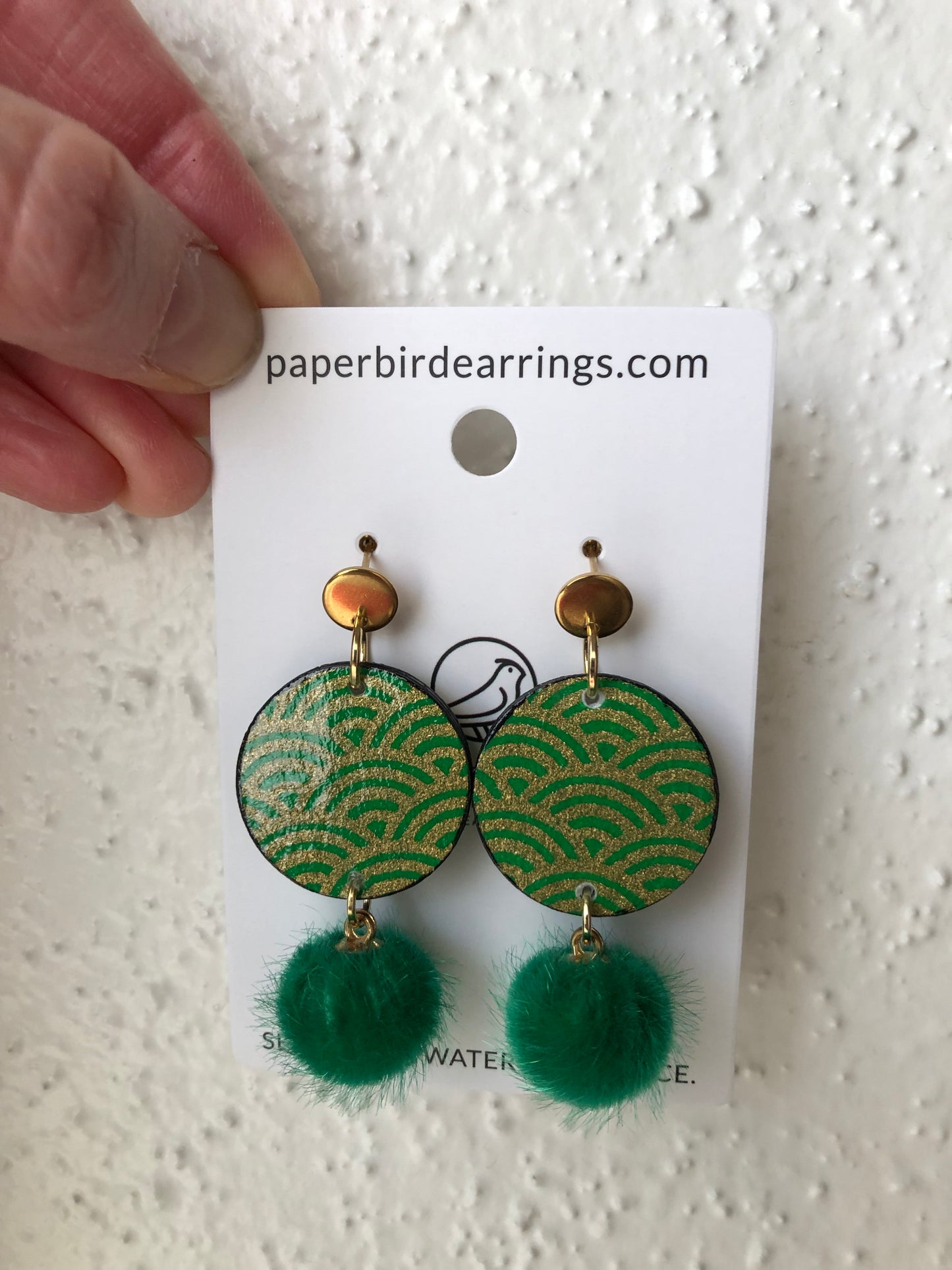 Green earrings with pom-poms on a paper card held by a hand.