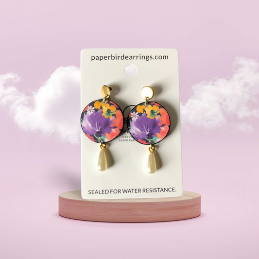 A pair of floral patterned round earrings with a gold-plated stainless-steel post and faux pearl detail, displayed on a cardboard stand with the text 'paperbird earrings.com' and 'sealed for water resistance'.