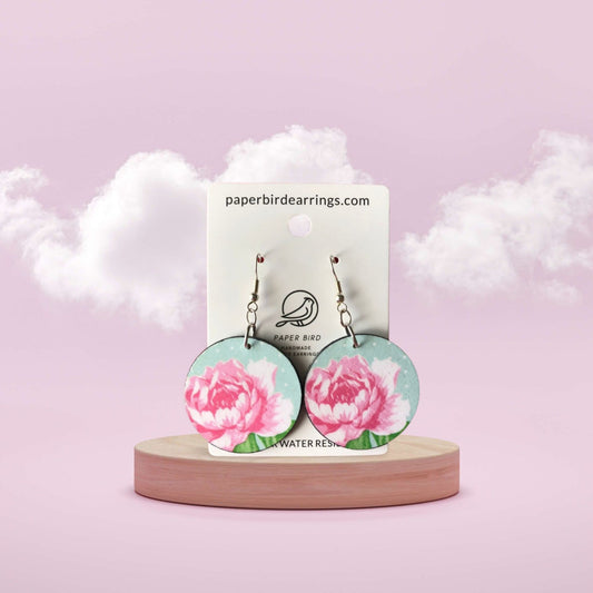 Floral earrings on a wooden stand with a branded card against a soft pink background