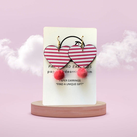 Heart-shaped earrings with pink stripes and red pom-poms on a white card with brand name, against a light purple background.
