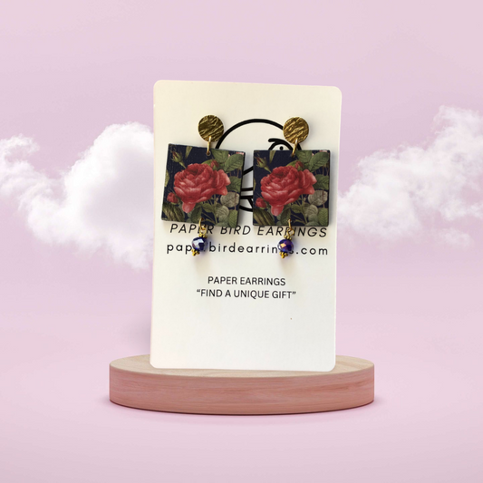 A pair of paper earrings with a rose flower design on a blue background, displayed on a wooden stand with the brand name 'PAPER BIRD EARRINGS' above.