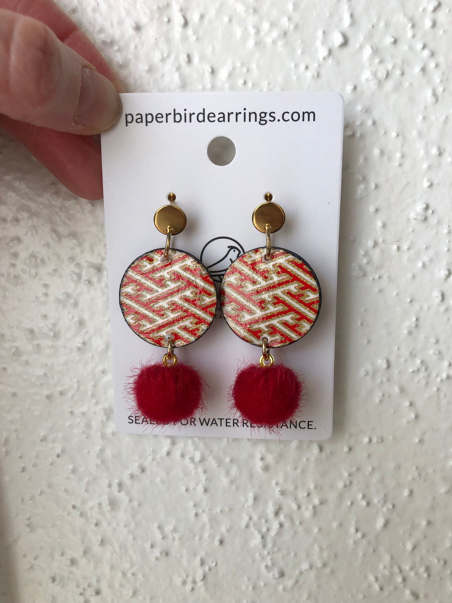 Pair of earrings with red pom-poms on a card with branding details.